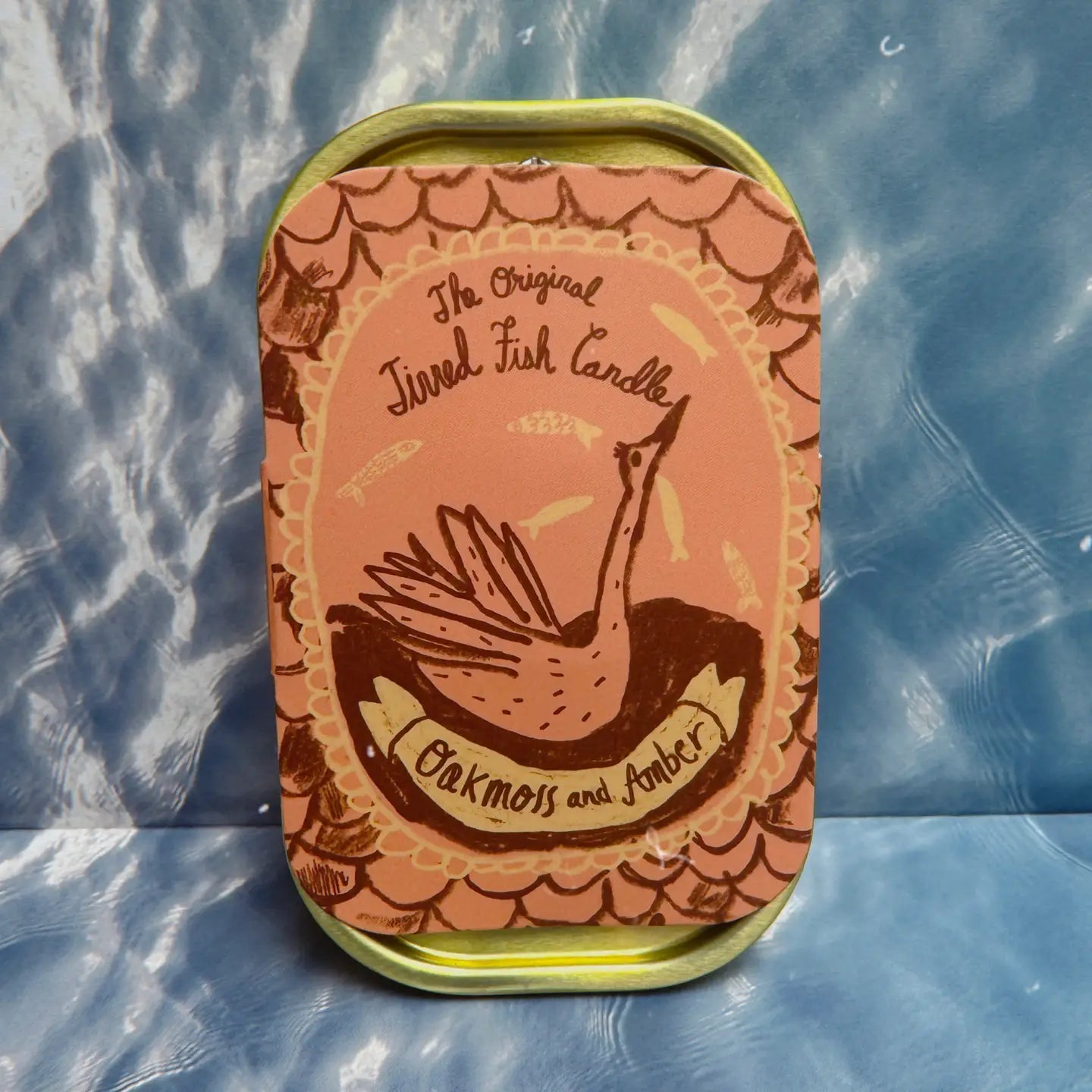 Rectangular tin with "The Original Tinned Fish Candle Oakmoss and Amber" label, winged fish illustration on pink-brown designs.