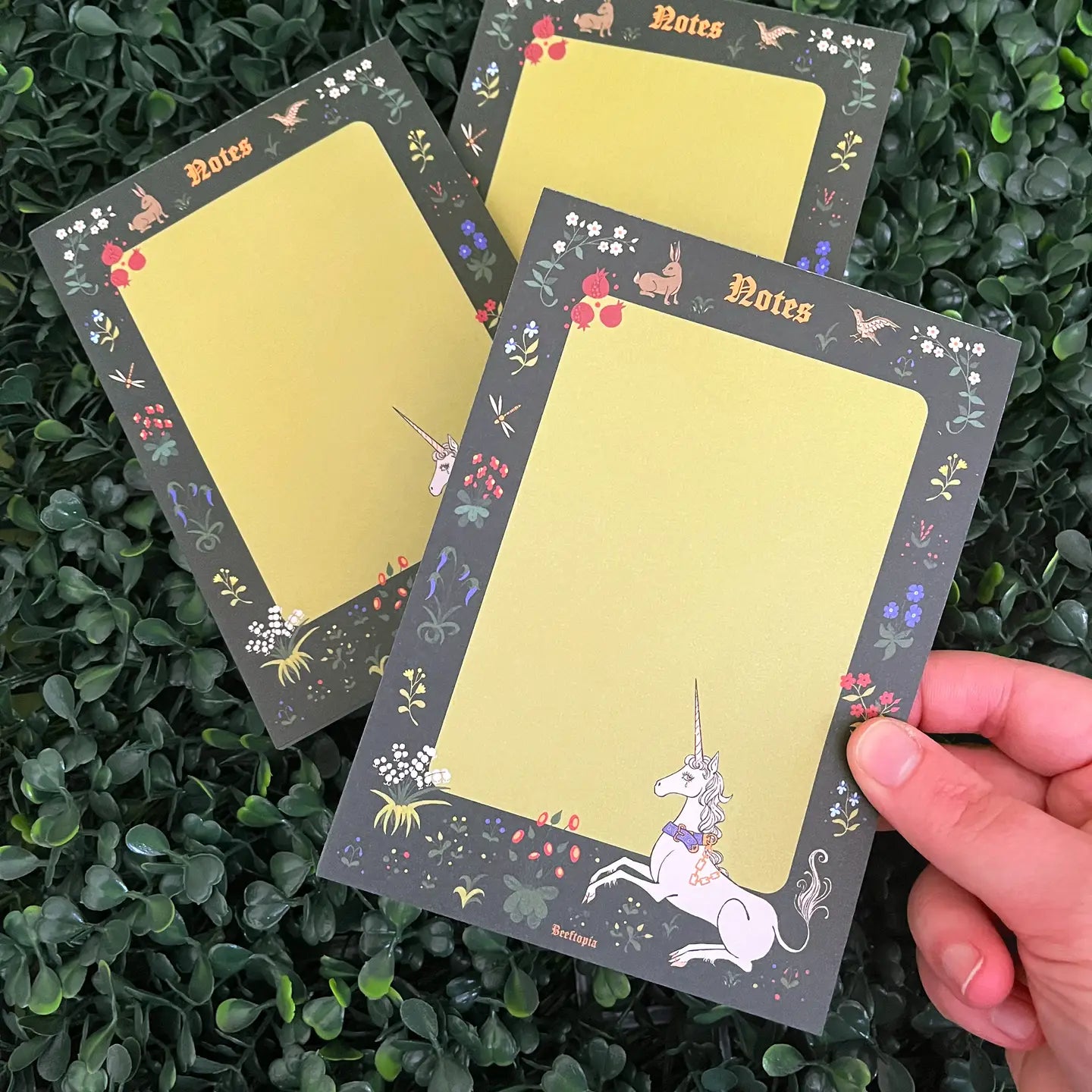 Hand holding Beeftopia Unicorn Tapestry A6 Notepad with floral cover, two notepads behind, on leafy background, medieval stationery.