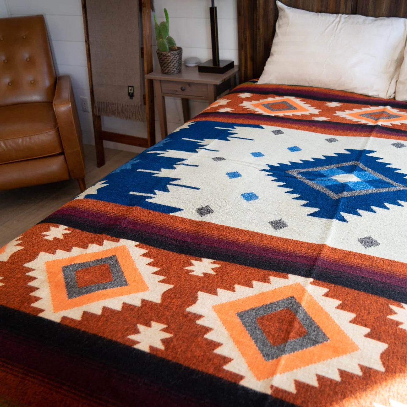 Alpaca Threadz Moab Orange Western Native Blanket on bed, near brown chair and nightstand. Cozy luxury rustic bedroom decor.