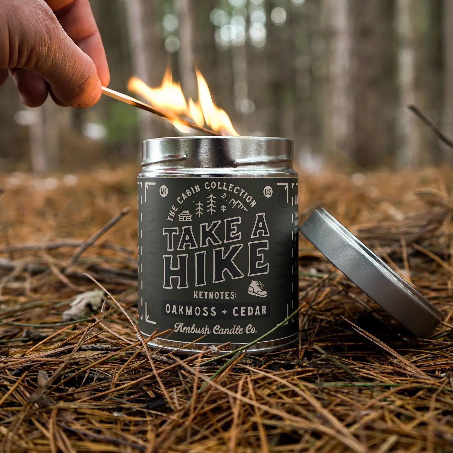 Hand lighting an eco-friendly Ambush Candle Co 14OZ Take A Hike Oakmoss & Cedar Candle in a forest setting, crafted with clean-burning soy wax. It perfectly complements your eco-friendly lifestyle.