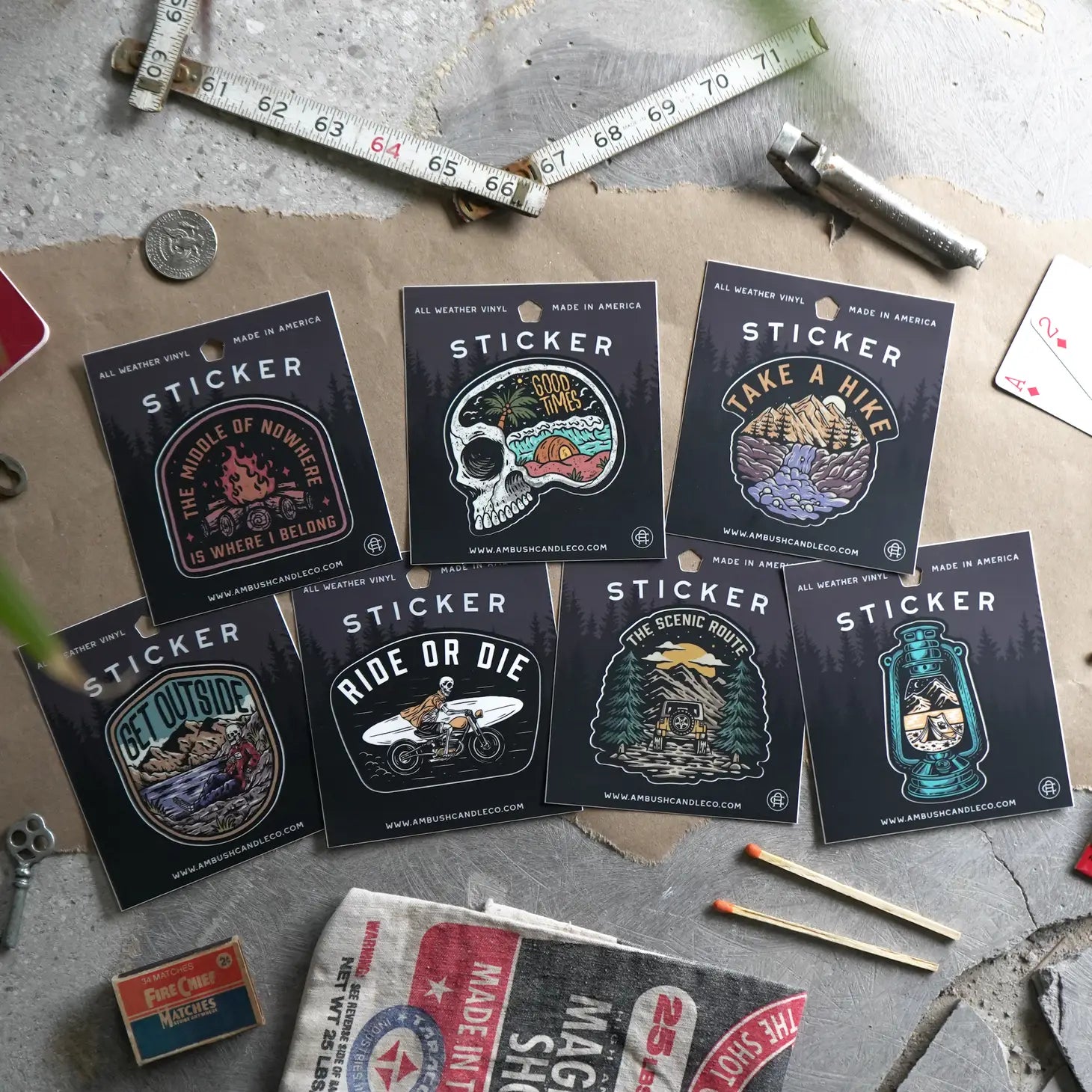 The Ambush Candle Co Take A Hike Sticker is a weatherproof vinyl sticker featuring "Take A Hike" text and a mountain scene, surrounded by vintage items like keys, matches, and playing cards. It's perfect for outdoor enthusiasts and hiker decal sticker collectors alike.