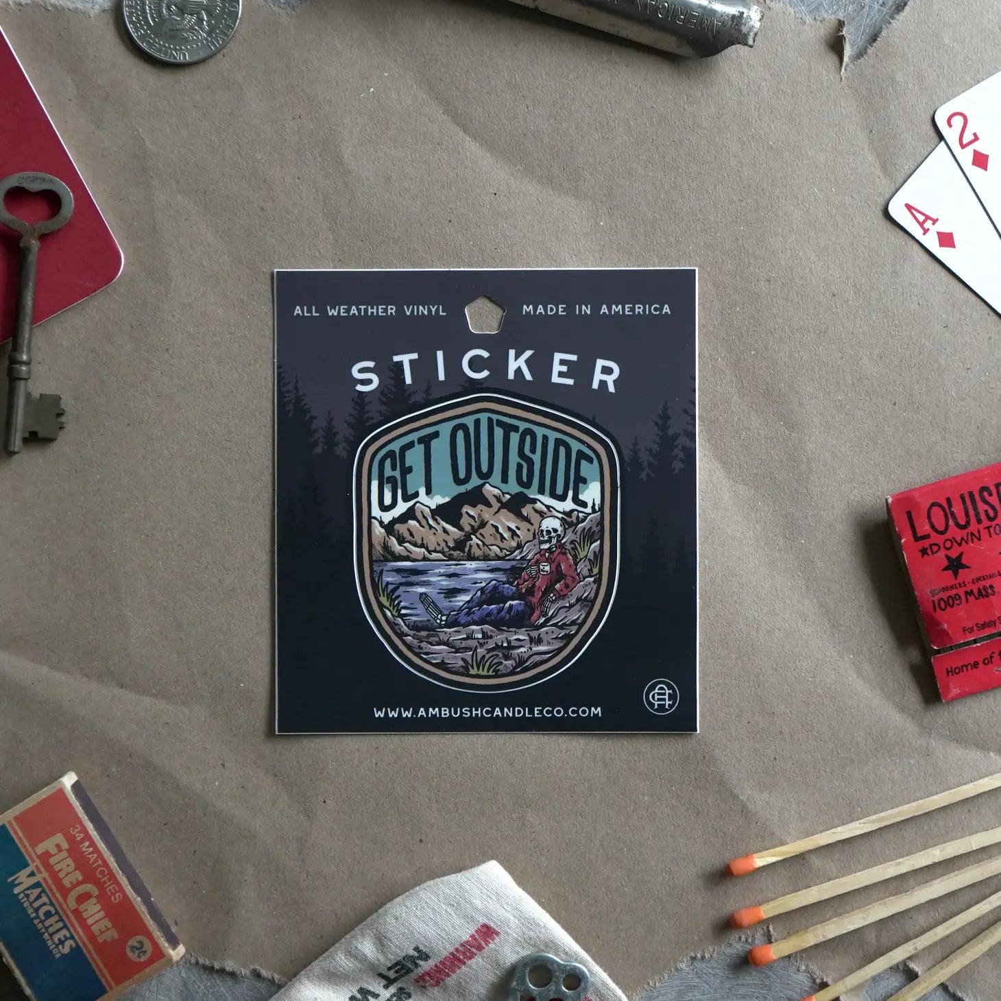 The Ambush Candle Co Get Outside Sticker, featuring the "GET OUTSIDE" text and a mountain scene, is placed among fallen autumn leaves. This weatherproof sticker, made in the USA, can be removed without damage.