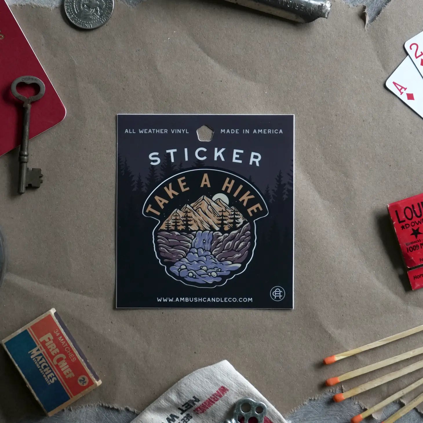 The Ambush Candle Co Take A Hike Sticker is a weatherproof vinyl sticker featuring "Take A Hike" text and a mountain scene, surrounded by vintage items like keys, matches, and playing cards. It's perfect for outdoor enthusiasts and hiker decal sticker collectors alike.