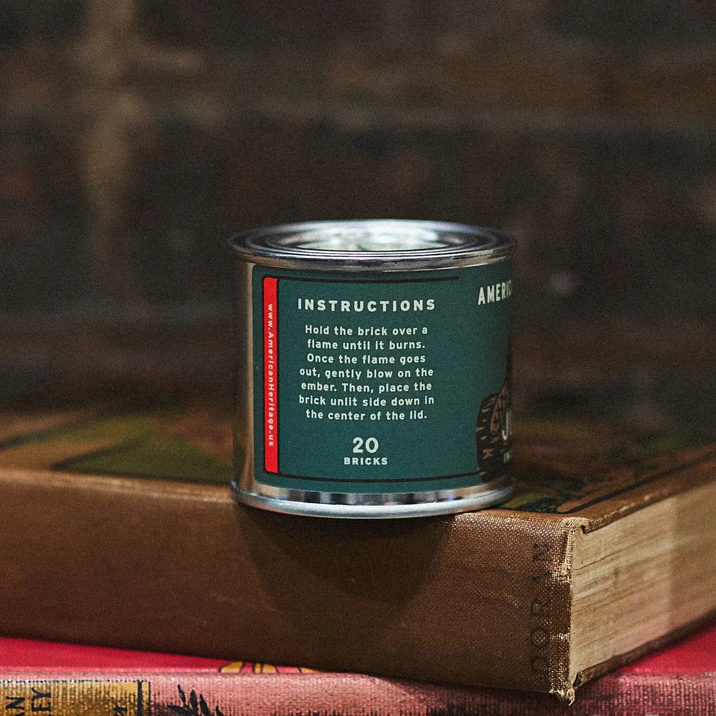 American Heritage Juniper 20 Incense Bricks metal tin rests on books, bringing a natural woodsy fragrance to your home décor.