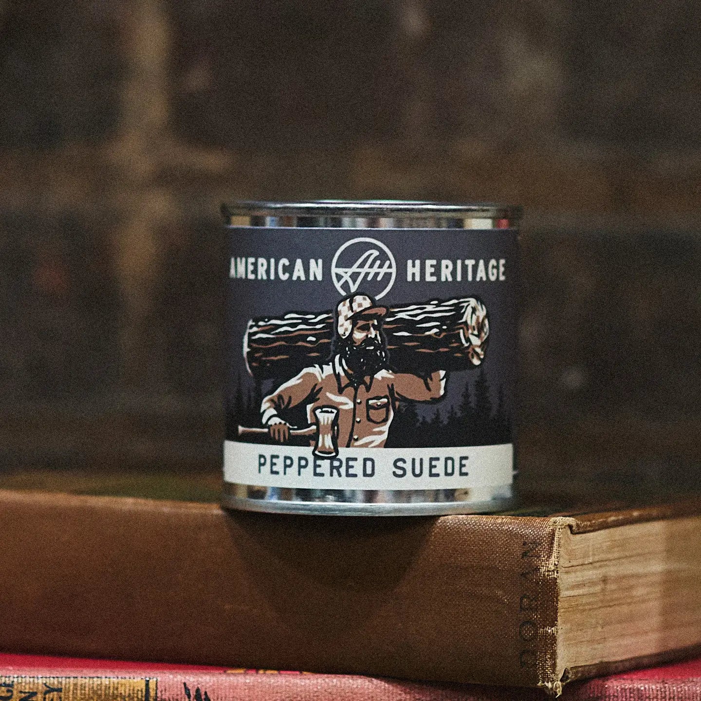 American Heritage Brands Peppered Suede Wood Wick Candle with lumberjack label sits atop stacked books, rustic home decor accent.