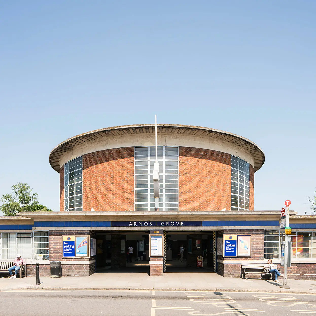 Arnos Grove station entrance, featuring semi-circular brickwork and large windows against a clear blue sky, stands as a testament to London Underground transport design; perfectly captured in the Blue Crow Media London Underground Architecture Map, with people seated on benches outside.