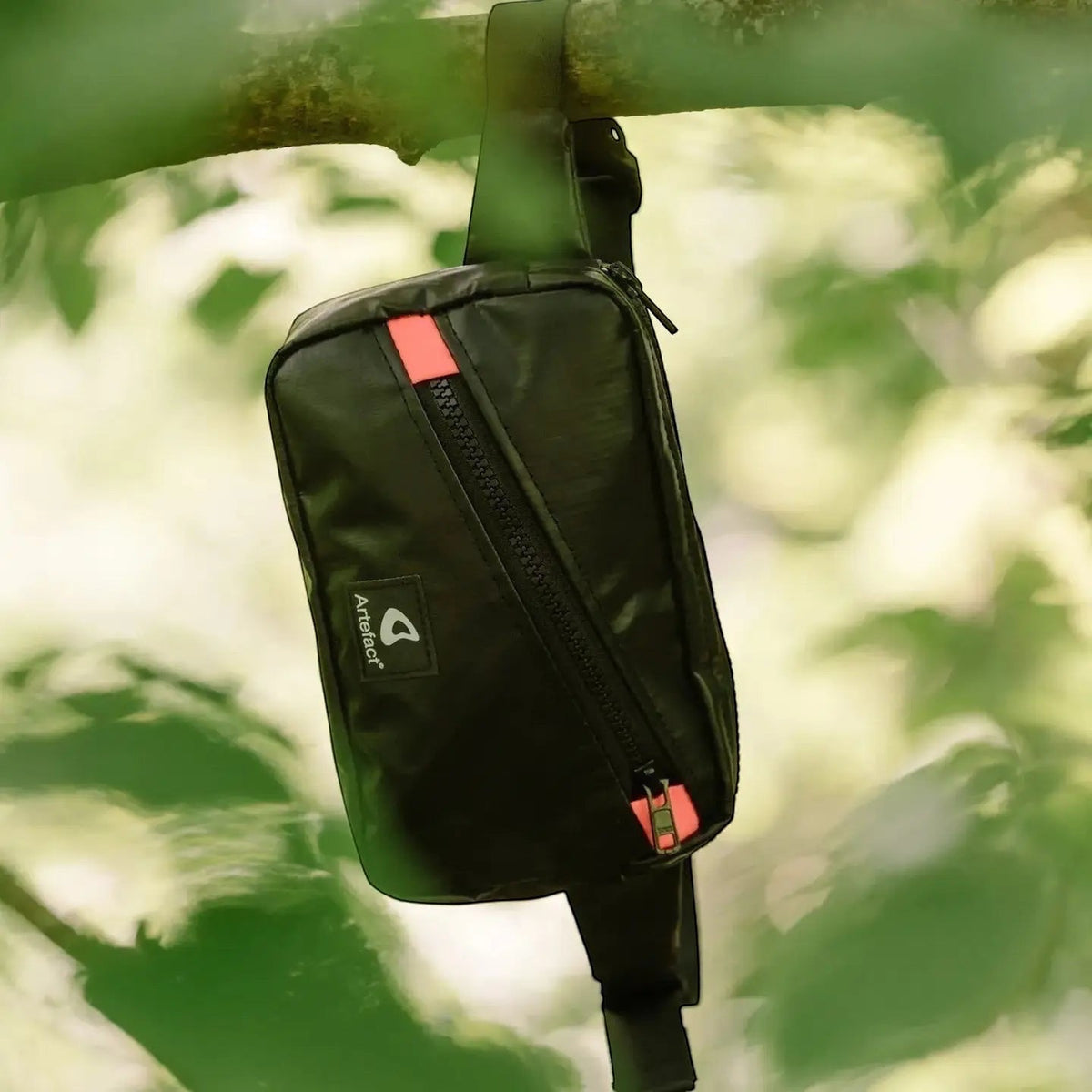 Black Artefact Eco Hornbill Banana Sling Bag hanging from a tree branch, surrounded by lush green leaves and pink accents.