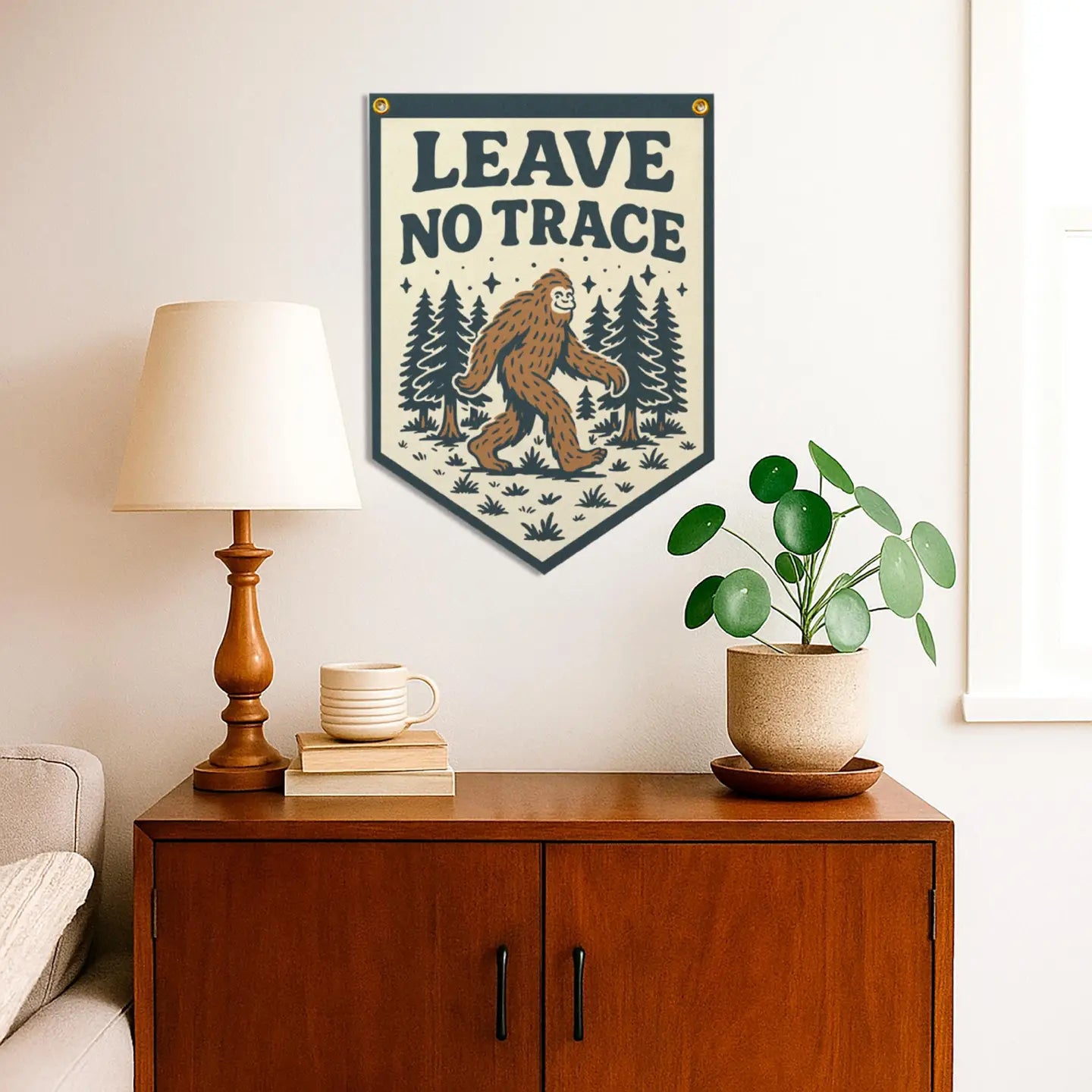A Benoit Leave No Trace Bigfoot banner pennant decorates a cabinet with a lamp, books, and plant—perfect nature cabin decor.