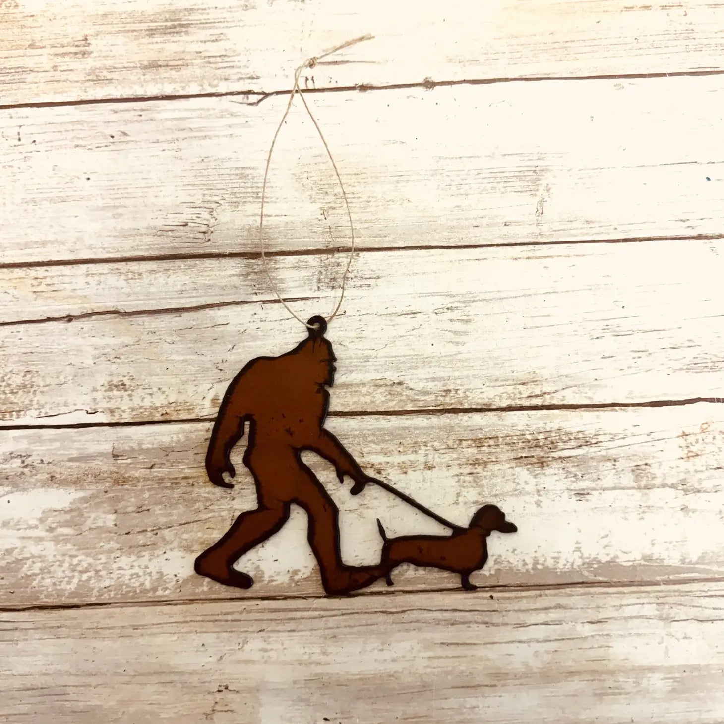 The Universal Iron Works Bigfoot Yeti Walking Sausage Dog Hanging Decoration, crafted from 18 gauge steel, is a metal silhouette of Bigfoot walking a sausage dog, displayed on a wooden background.