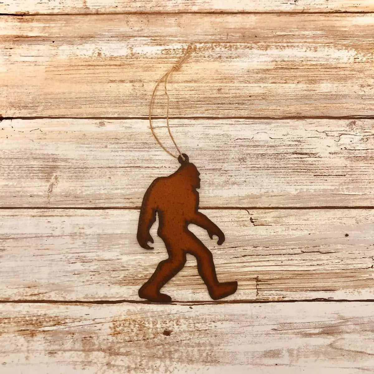 Presenting the Universal Iron Works Bigfoot Walking Hanging Decoration, a striking silhouette of Bigfoot crafted from 18 gauge steel. This ornament, complete with a looped string for easy hanging, stands out beautifully against its rustic wooden background. The natural rusting process imparts an authentic touch to this distinctive piece.