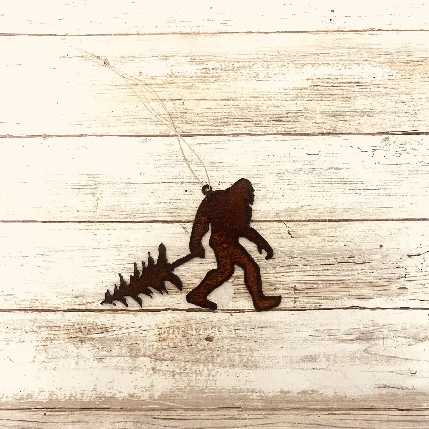 The Universal Iron Works Bigfoot Yeti With Tree Hanging Decoration, crafted from 18 gauge steel and shaped like the mythical creature carrying a Christmas tree, hangs against a weathered wooden background. The natural rusting process gives it a rustic charm, perfect for holiday decor.