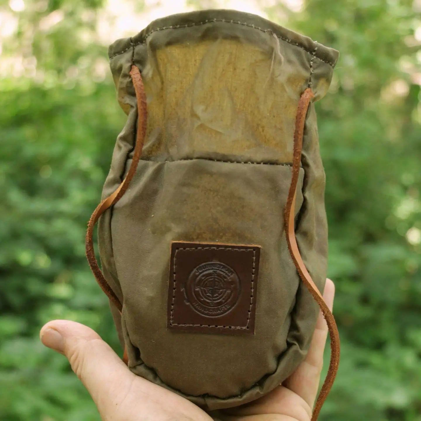 Hand holding Journeyman Craft Khaki Green Waxed Canvas Coffee Pouch outdoors among lush greenery, travel gear for coffee lovers.