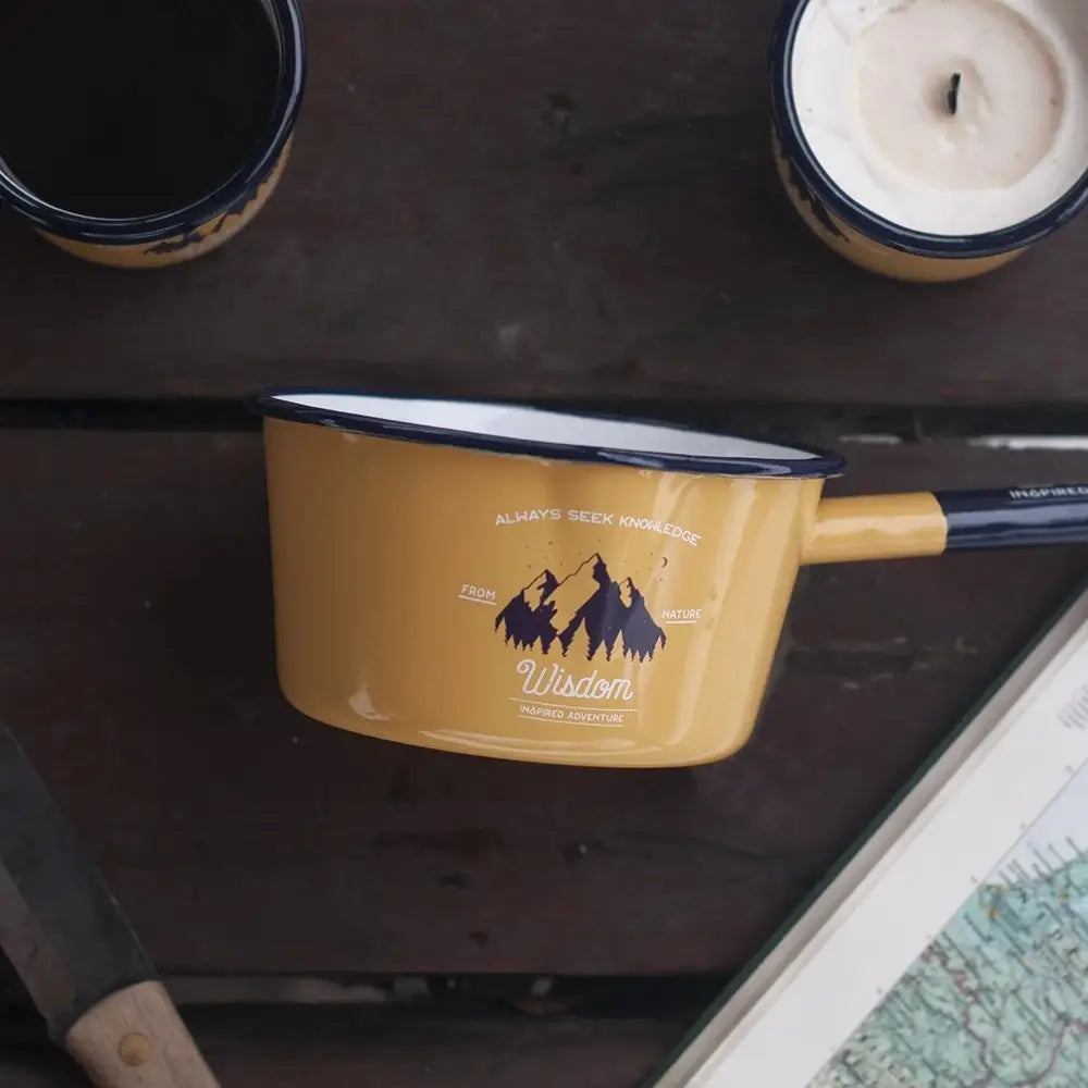 The Emalco Enamelware Enamel Wisdom Saucepan, sporting a vibrant yellow hue with a mountain design and the word "Wisdom," rests on a wooden surface beside a candle, map, and knife.