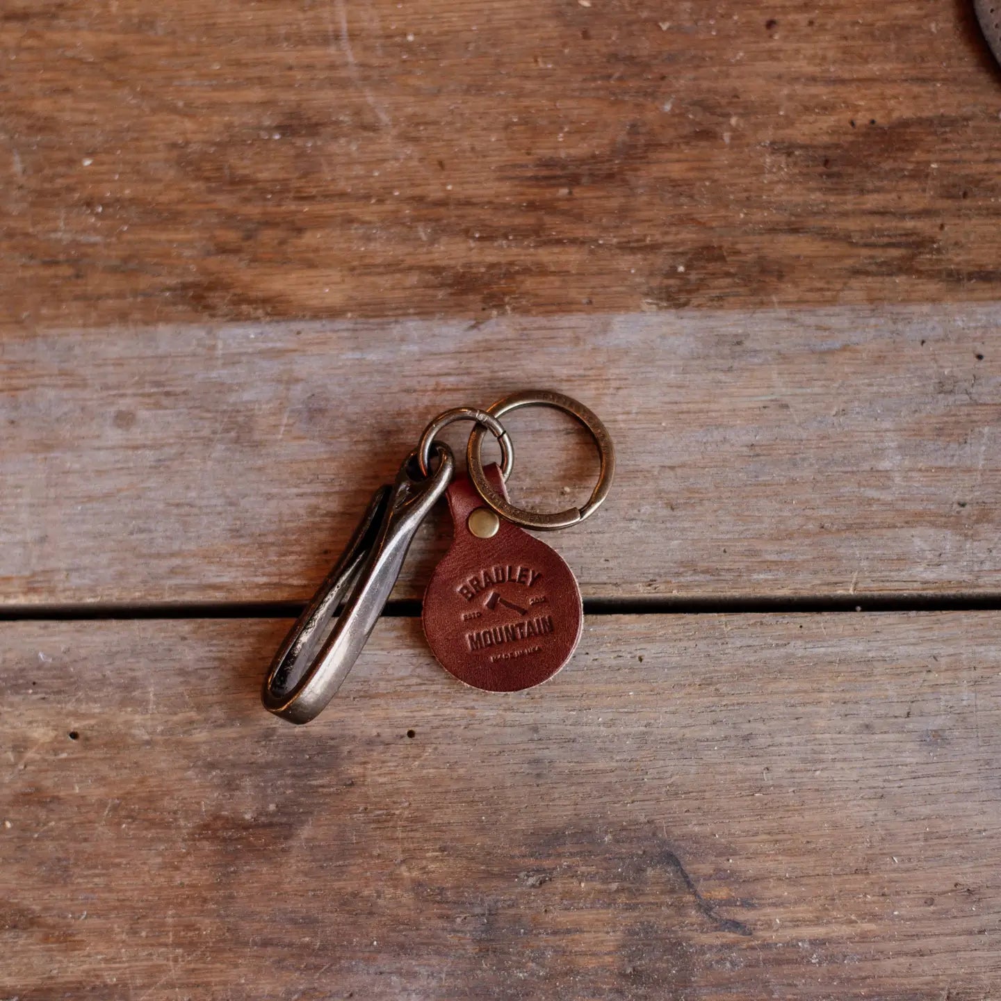 Bradley Mountain Brass Fish Hook Key Fob, brown leather tag, and carabiner displayed on rustic wooden surface for sale.