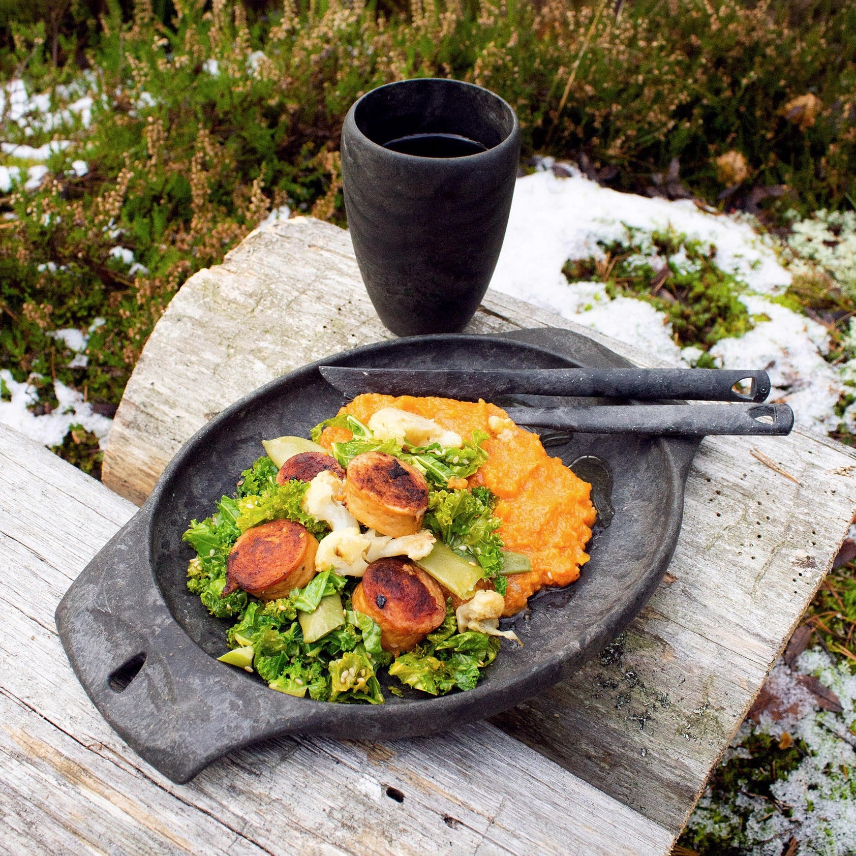 Black plate with greens, cheese & patties next to Kupilka Cutlery Set and black cup on wooden bench—camp meal outdoors.