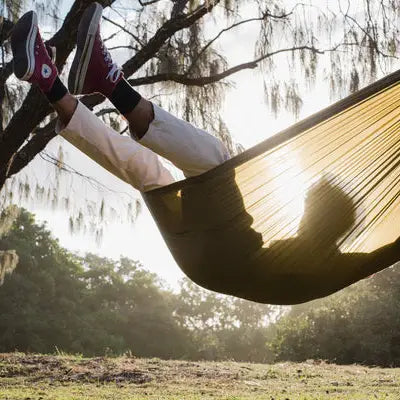 Enjoy ultimate relaxation with legs raised in the Ghost Outdoors Peacock Hammock, surrounded by sunlit trees and lush green grass.
