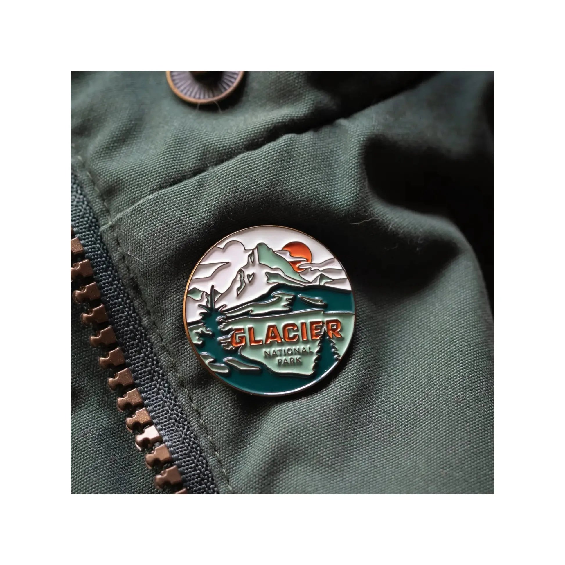 The Landmark Project Glacier National Park Pin is a round enamel pin featuring a mountain and river design with the text "GLACIER NATIONAL PARK." Attached to a green fabric backing, this pin celebrates Glacier National Park and highlights our dedication to sustainable packaging, making it an ideal addition for enamel pin collectors.
