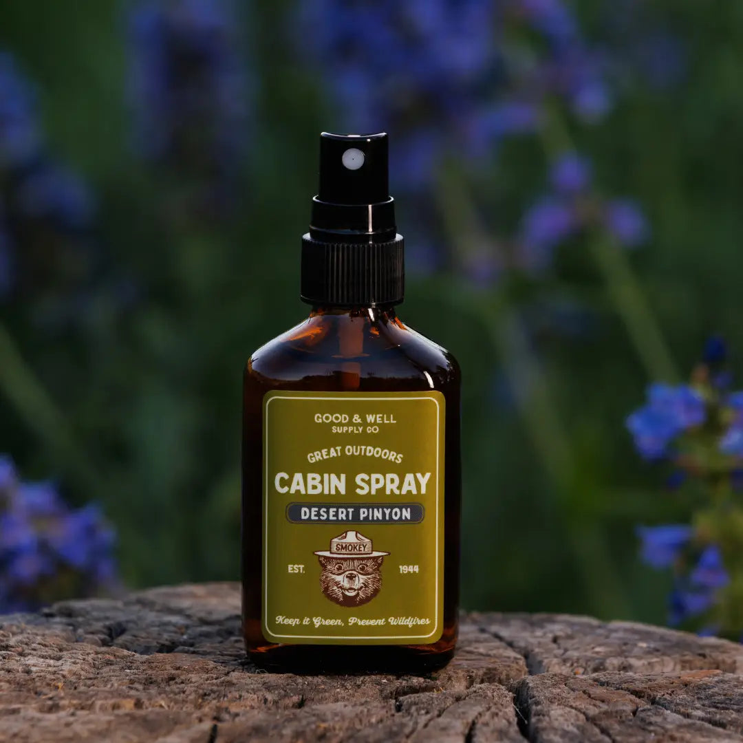Good & Well Supply Co Desert Pinyon Smokey Bear Cabin Spray bottle on a rock, purple wildflowers in the background.