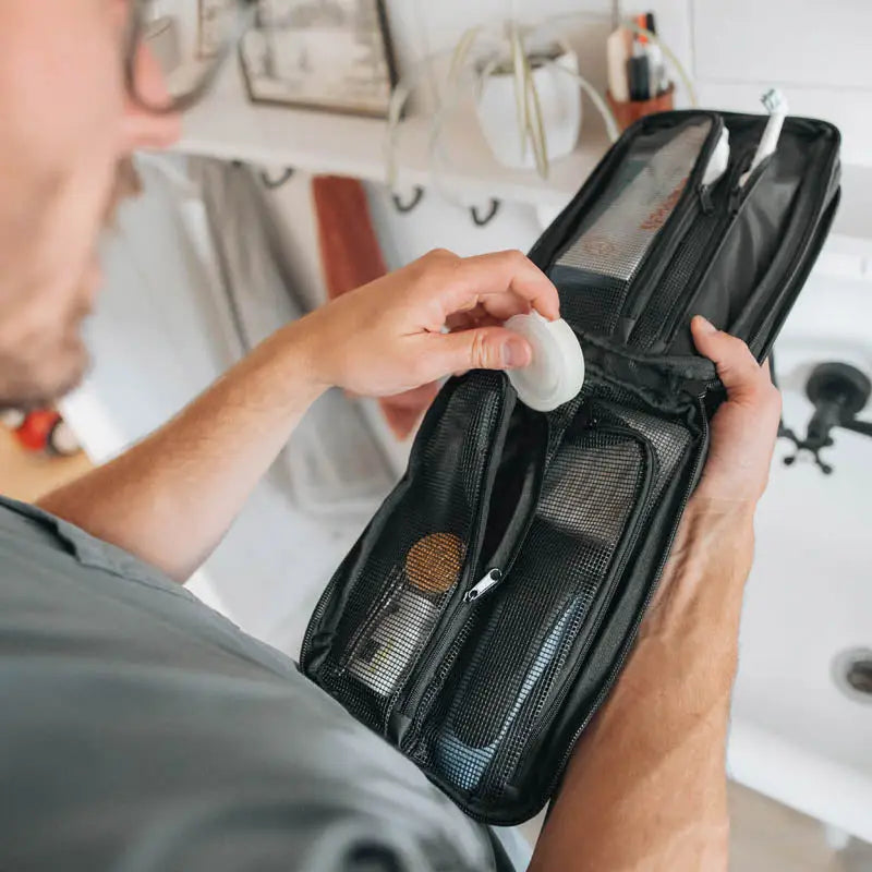 A person packs a round item into the Gravel Outdoor Black Explorer Plus™ Toiletry Bag with organized zippered compartments.