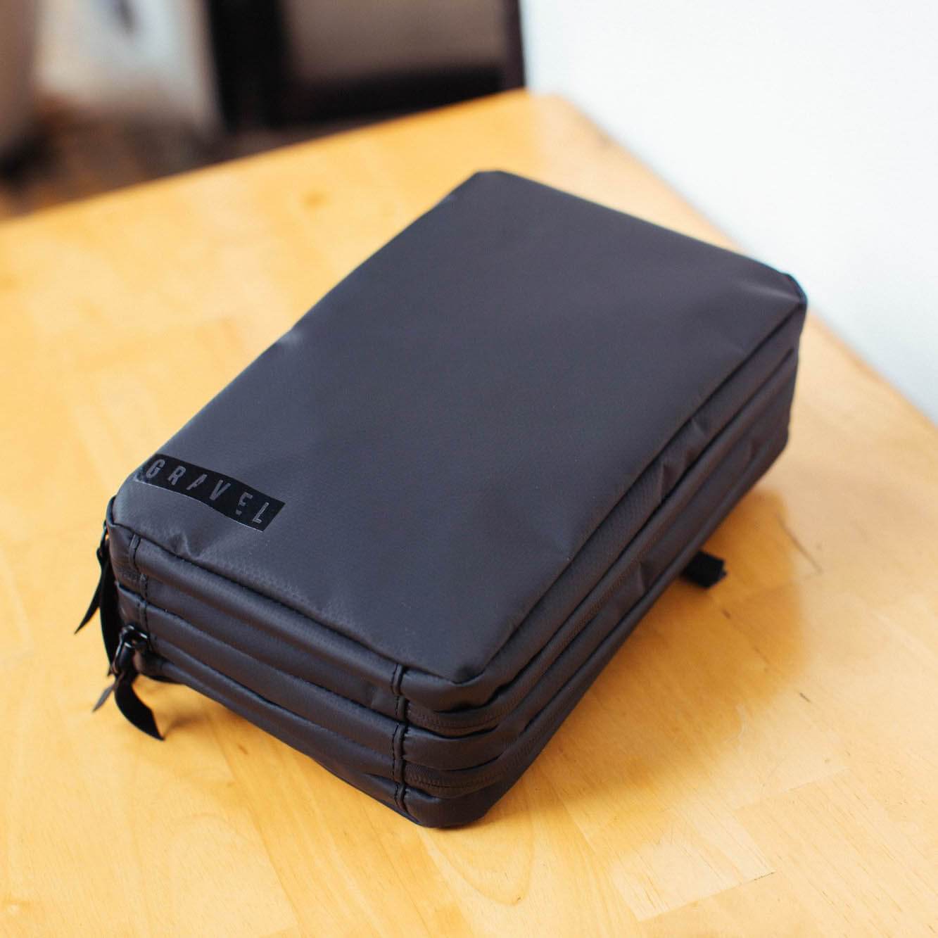 Gravel Explorer Plus™ black toiletry bag closed on a light wooden table—waterproof, travel organizer, mens grooming accessory.
