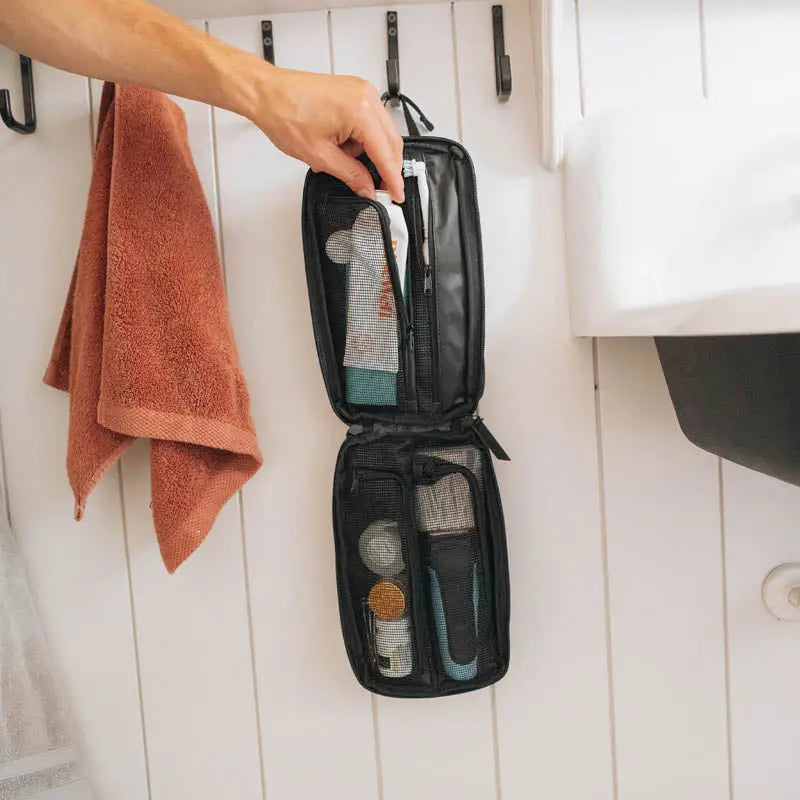 Hand holds Gravel Explorer Plus™ TSA-approved black toiletry bag, hanging on bathroom hook—durable travel organizer pouch.