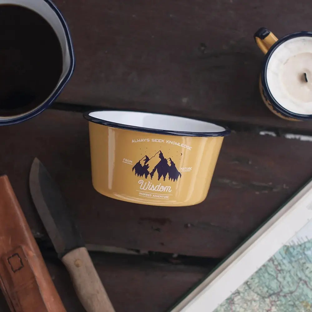 The Emalco Enamelware 25.2oz Enamel Wisdom Bowl is a yellow enamel bowl with a blue rim, adorned with mountain designs and the inspirational texts "Always Seek Knowledge," "From Nature," and "Wisdom Inspired Adventure." This Wisdom Bowl is not only motivational but also crafted from durable material.