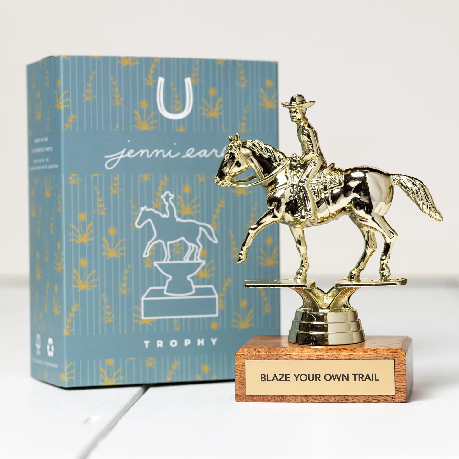 The Jenni Earle Cowboy Trophy - Blaze Your Own Trial features a stunning golden horse and rider on an FSC-certified wooden base with the inspiring text "BLAZE YOUR OWN TRAIL". It comes in a decorative, recyclable gift box adorned with matching imagery, making it an ideal trophy gift for any trailblazer.