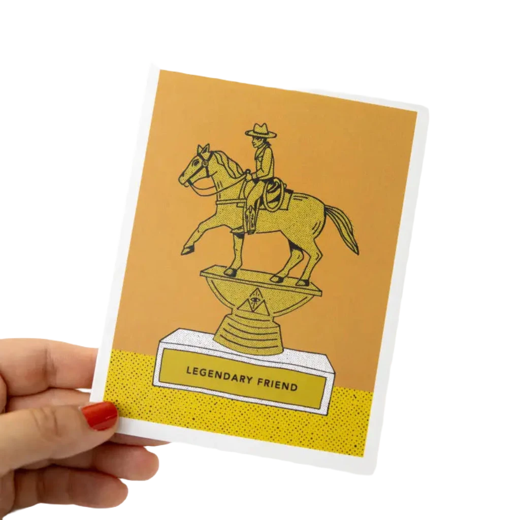 A hand holds the Jenni Earle Legendary Friend Greeting Card, meticulously hand-illustrated in a Wes Anderson western style. The card features a cowboy riding a horse on a trophy base labeled "Legendary Friend," crafted from 100% recycled paper.