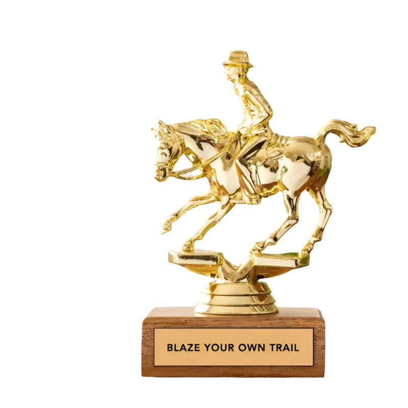 The Jenni Earle Cowboy Trophy - Blaze Your Own Trial features a stunning golden horse and rider on an FSC-certified wooden base with the inspiring text "BLAZE YOUR OWN TRAIL". It comes in a decorative, recyclable gift box adorned with matching imagery, making it an ideal trophy gift for any trailblazer.