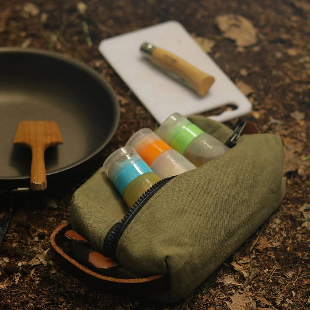Forager Provisions LTD green canvas toiletry bag filled with travel bottles, sitting on forest ground near camping gear.
