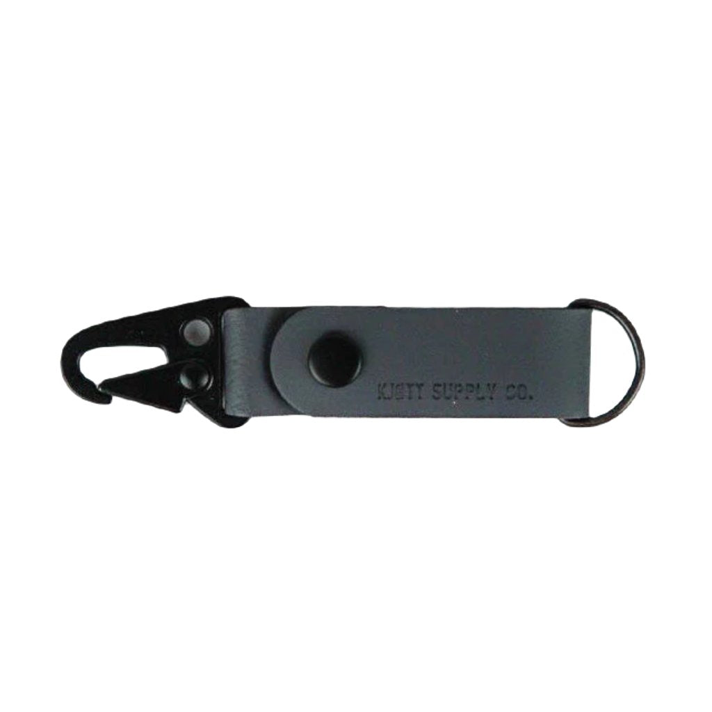 A blue Kjøtt Supply Co Key Fob, featuring a black metal hook, a black button, and the label "KOTT SUPPLY CO." against a white background, crafted from premium leather and backed by a lifetime warranty.