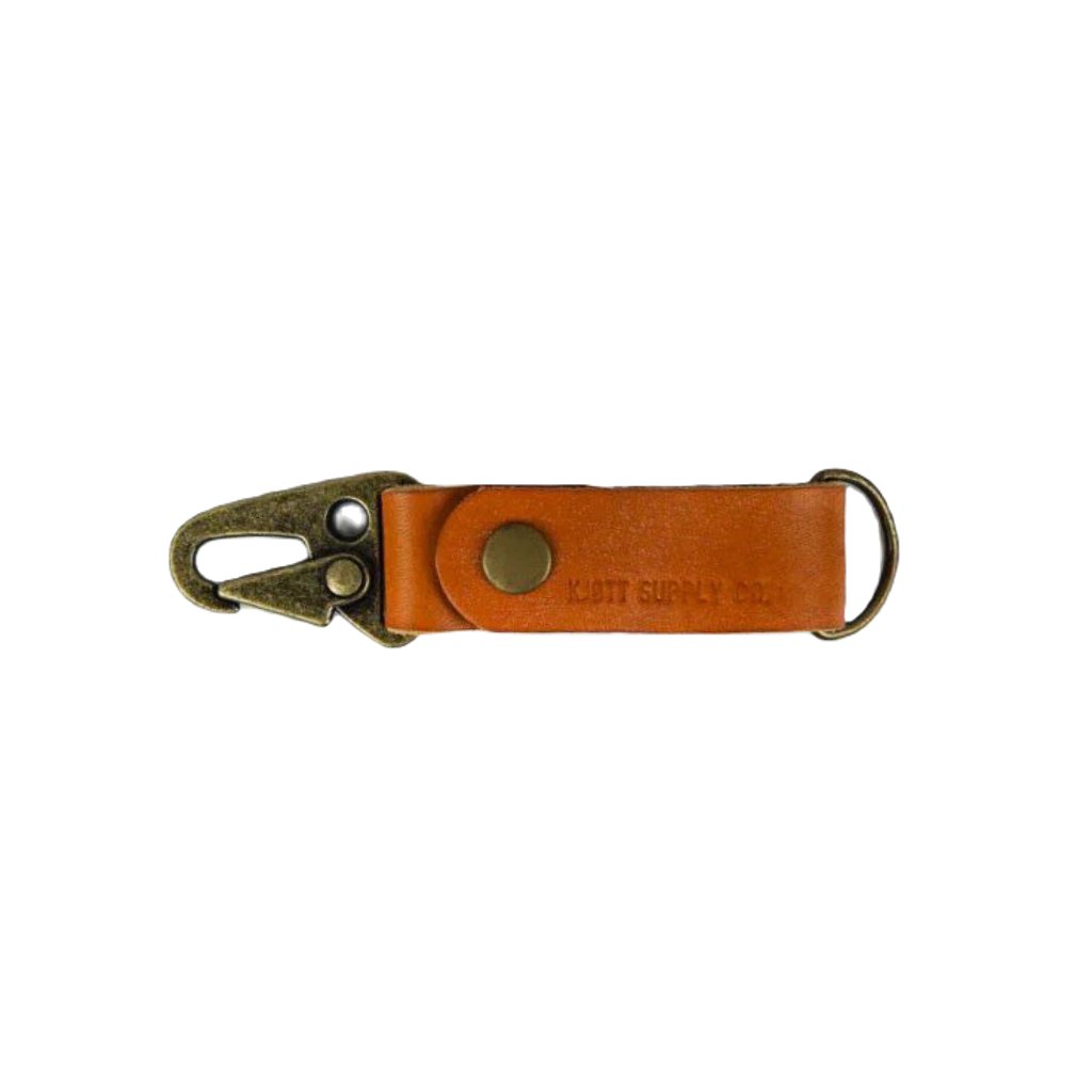 A blue Kjøtt Supply Co Key Fob, featuring a black metal hook, a black button, and the label "KOTT SUPPLY CO." against a white background, crafted from premium leather and backed by a lifetime warranty.