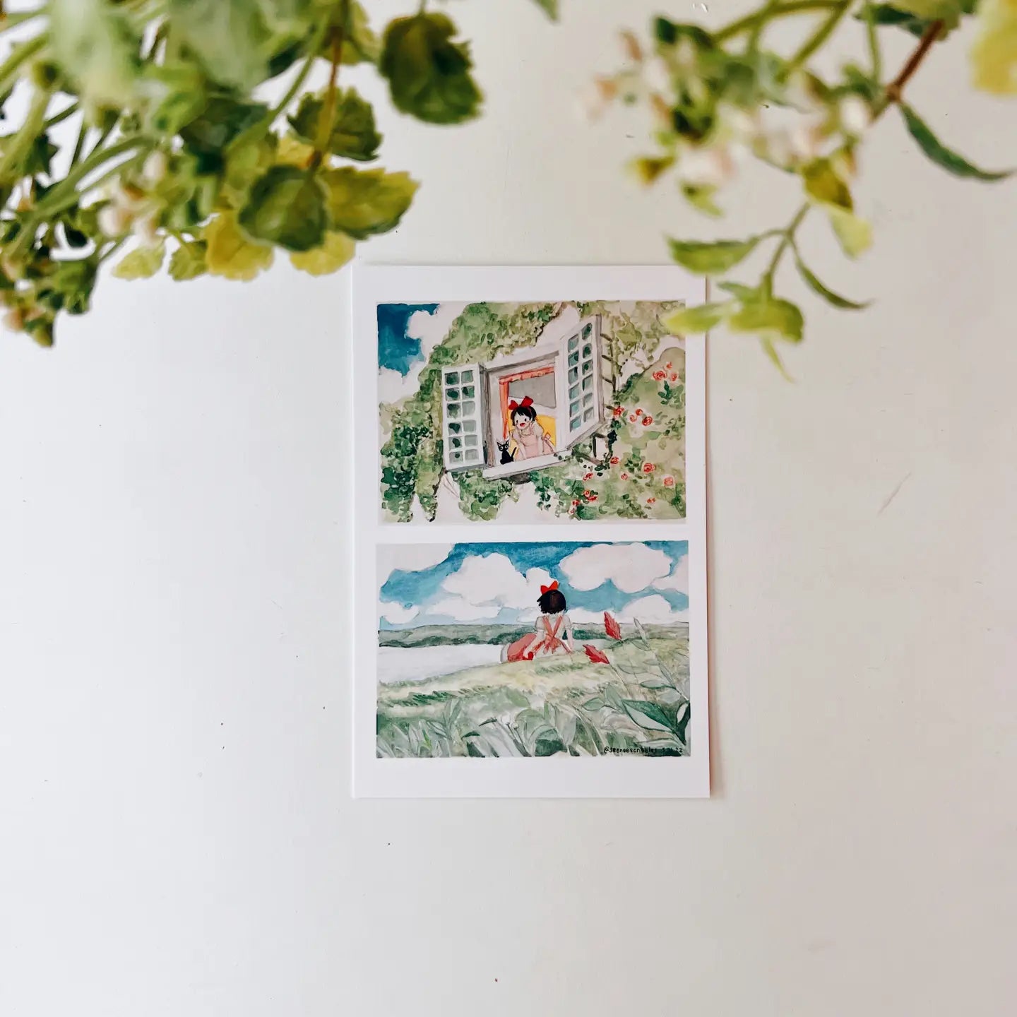 A hand holds the Seenee Scribbles Art Print 4 x 6 - Kiki On A Clear Day, featuring two beautiful watercolor illustrations: a girl at a flowered window (top) and a girl in a field with birds (bottom), evoking scenes from Kiki's Delivery Service.