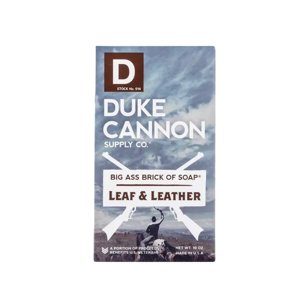 The packaging of Duke Cannon Big Ass Brick Of Soap Leaf & Leather features a rugged outdoor background that underscores its masculine scent and substantial size while also emphasizing its American-made quality and benefits for veterans.