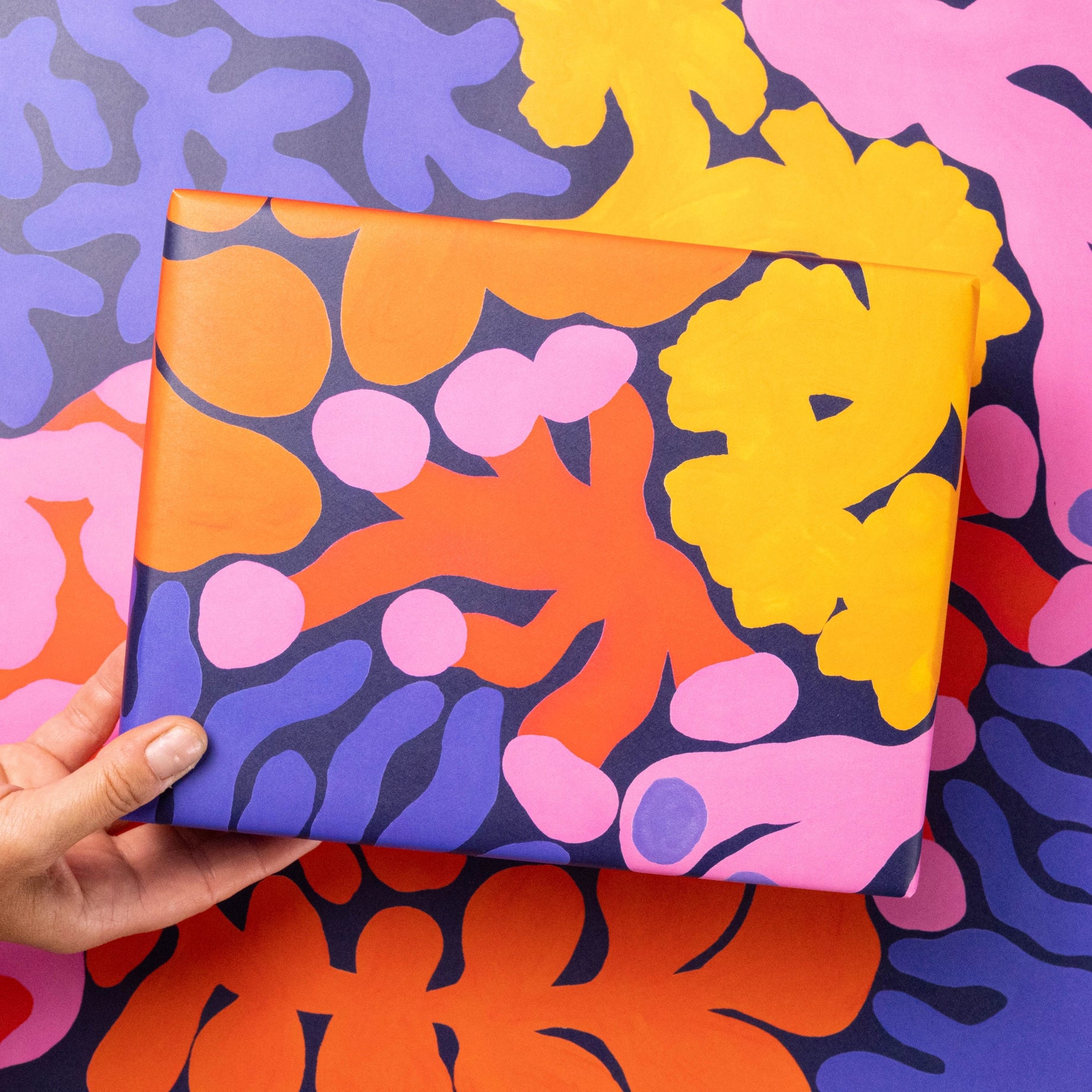 Hand holding Evermade Coral Gift Wrap by Liv Lee X WWF featuring vibrant abstract shapes in yellow, orange, pink, and purple.