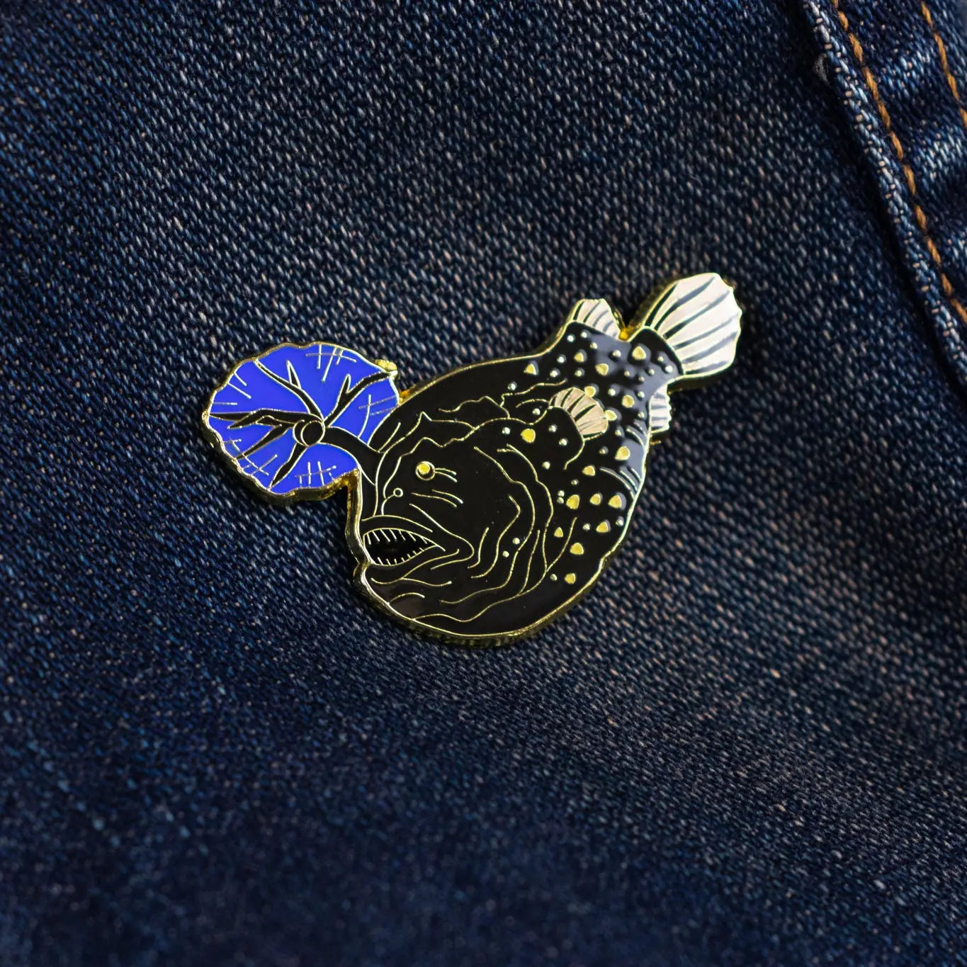 Stemcell Atlantic Football Fish enamel pin with blue lure displayed on dark blue denim. Unique ocean-themed collectible pin.