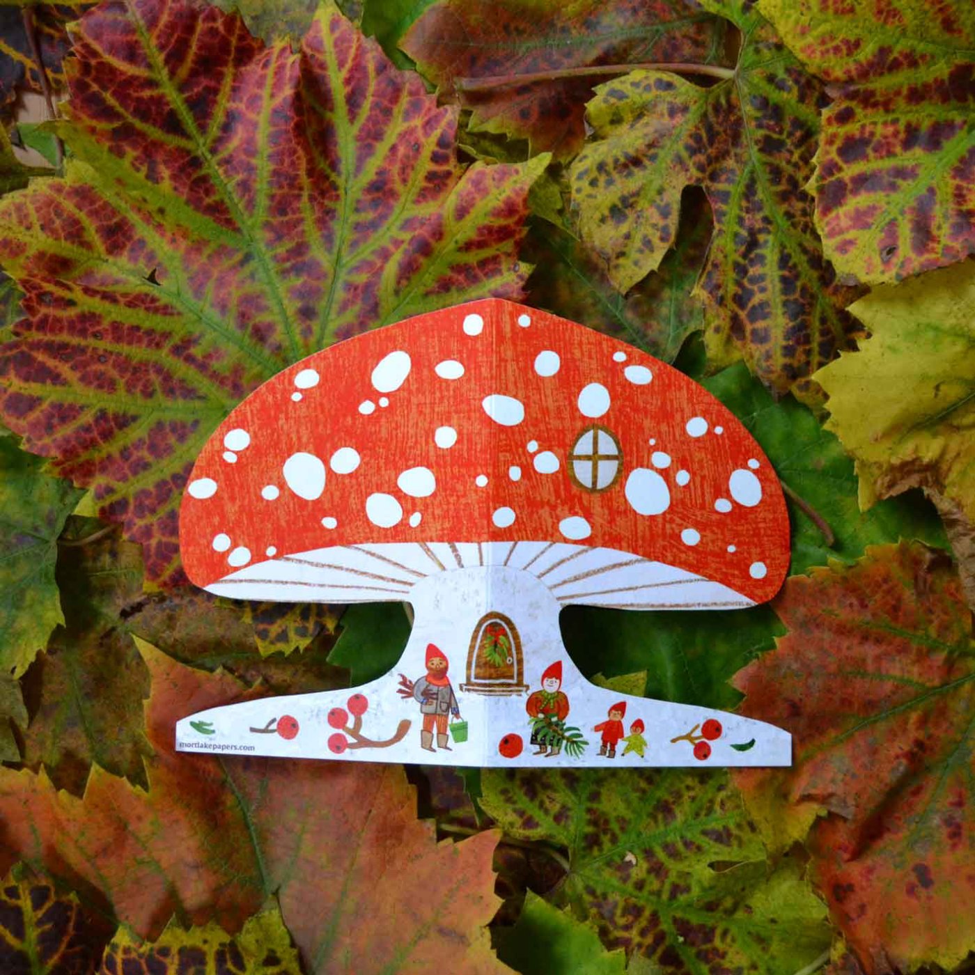 Mushroom shaped card featuring gnomes collecting foliage laid out on autumnal coloured leads.