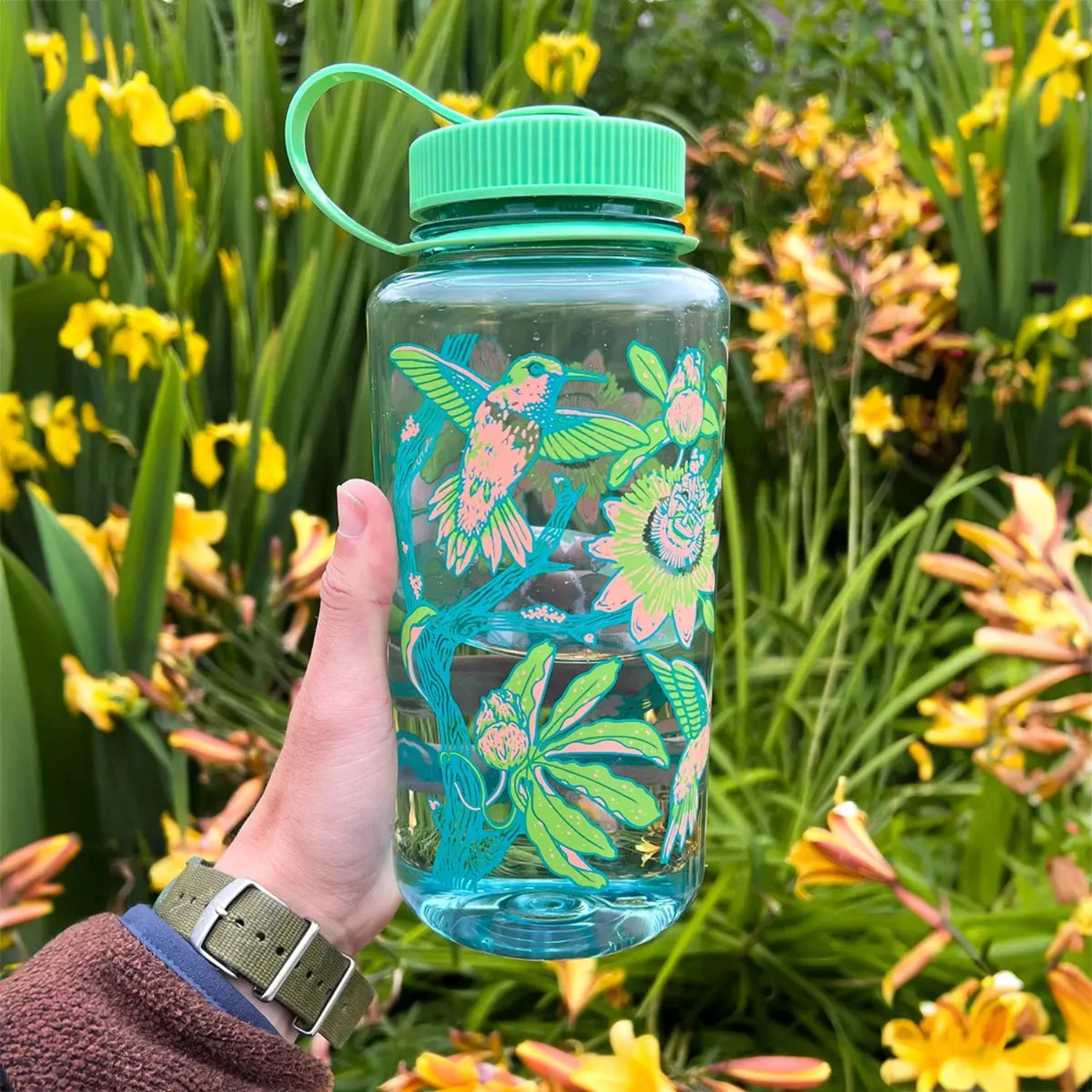 Hand holding Mustard Beetle 32oz Wide Mouth Nalgene Hummingbird Water Bottle with green lid, outdoors in yellow wildflowers.