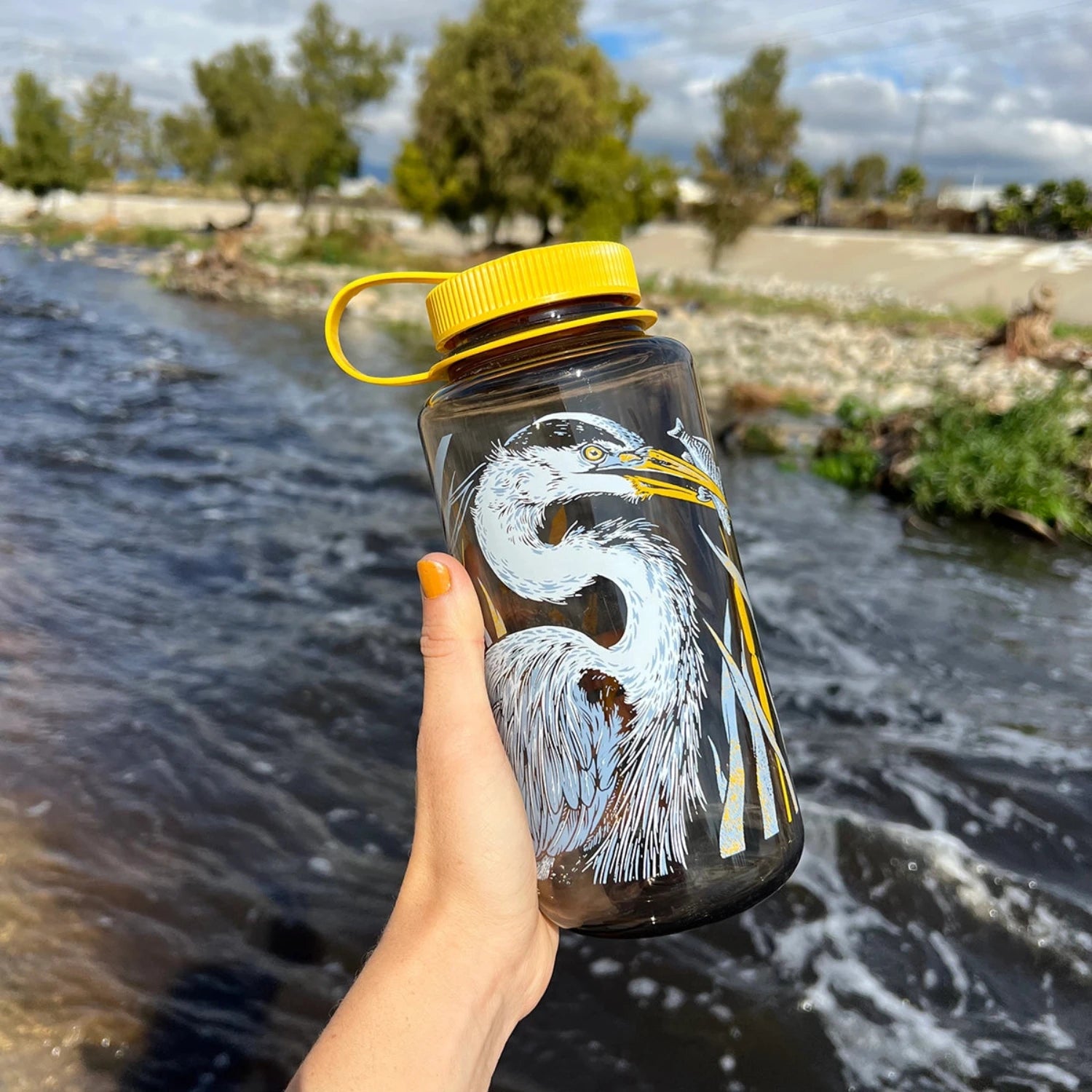 Hand holding a Mustard Beetle 32oz Nalgene Pasadena Audubon LA River edition bottle by the river, eco-friendly water bottle.