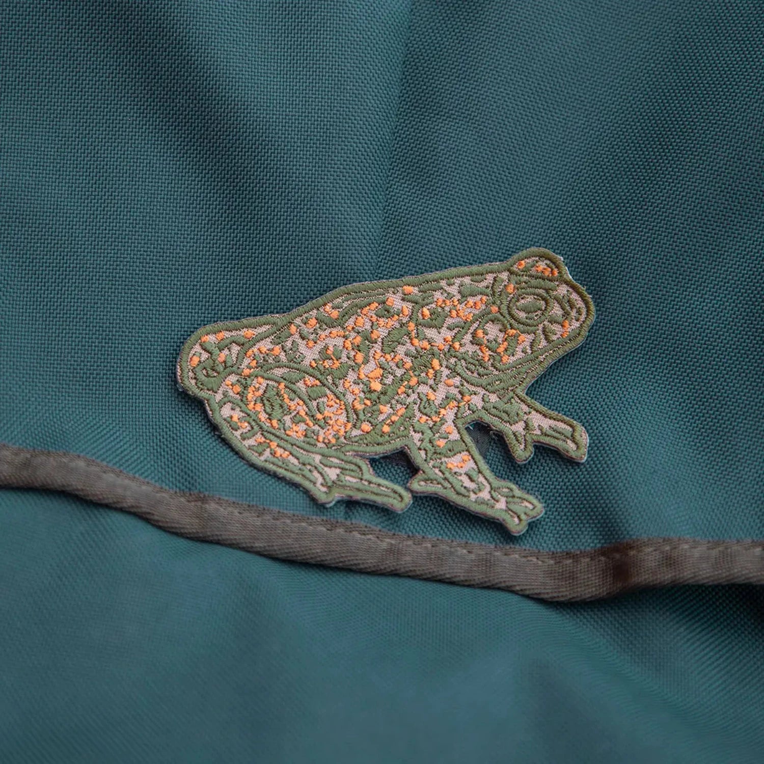 Mustard beetle toad iron-on patch sewn on teal fabric, with a bold seam underneath; unique embroidered applique detail.