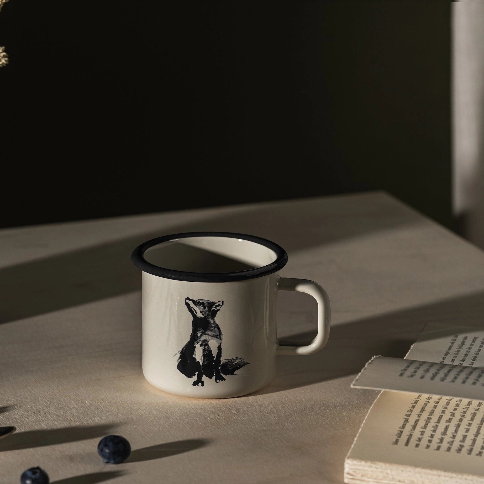 Muurla Teemu Järvi 0.37L Enamel Mug with Fox sits on table beside open book, fresh blueberries scattered, cozy kitchen scene.