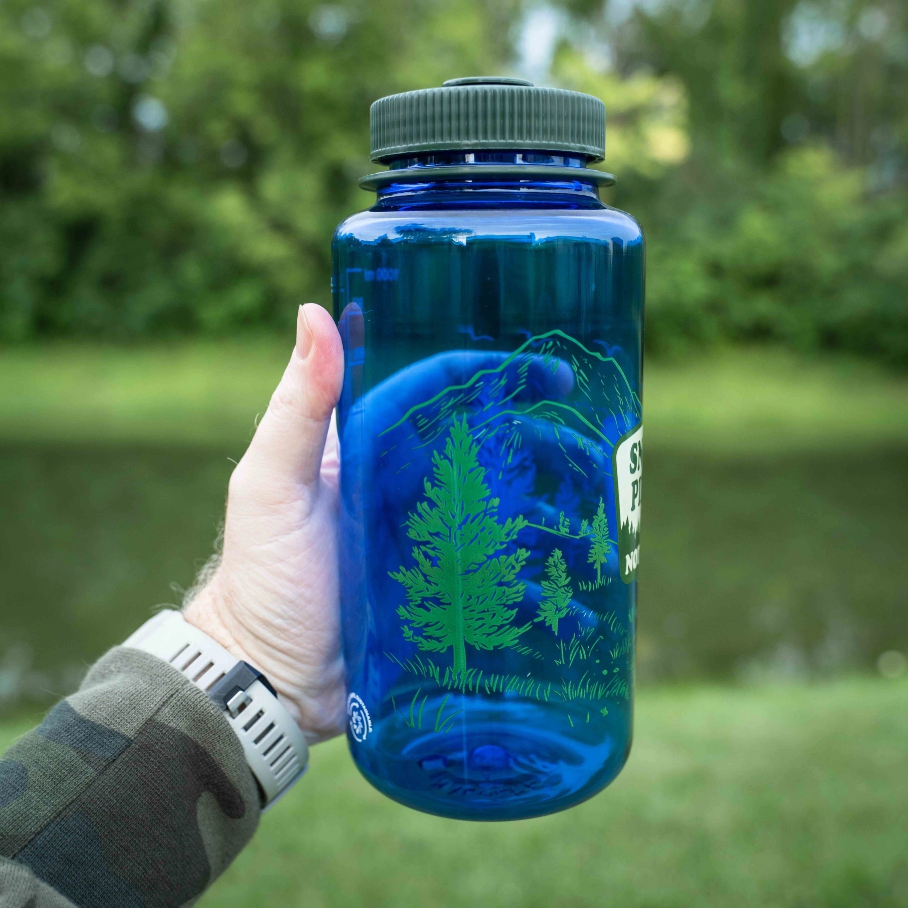 Forager Provisions LTD blue Nalgene 32oz water bottle with Snort Pines Not Lines graphic held outdoors near a lake