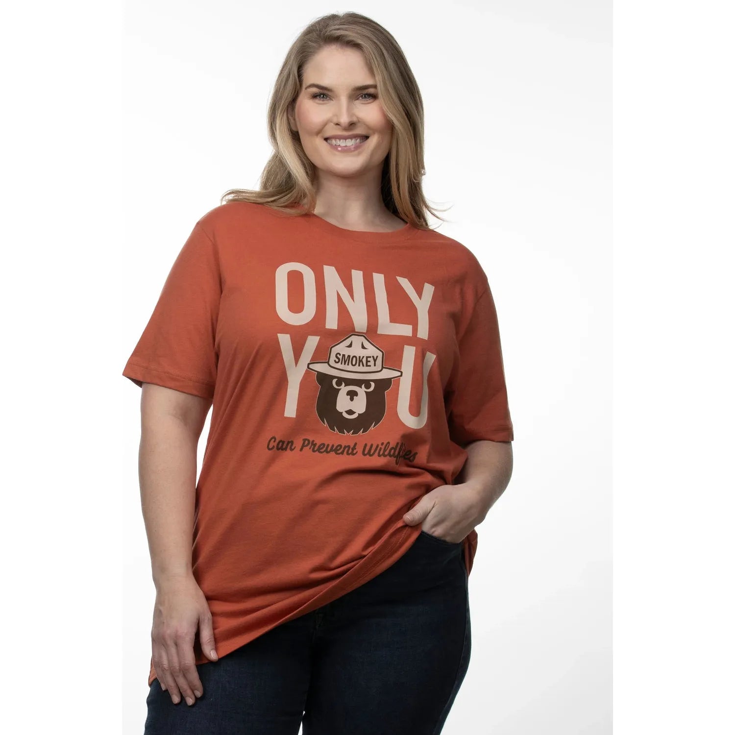 A woman smiles, wearing The Landmark Project Only You Smokey Bear T-Shirt in Spice with the text "Only You Can Prevent Wildfires," against a plain background. This sustainably made shirt underscores the importance of wildfire prevention.
