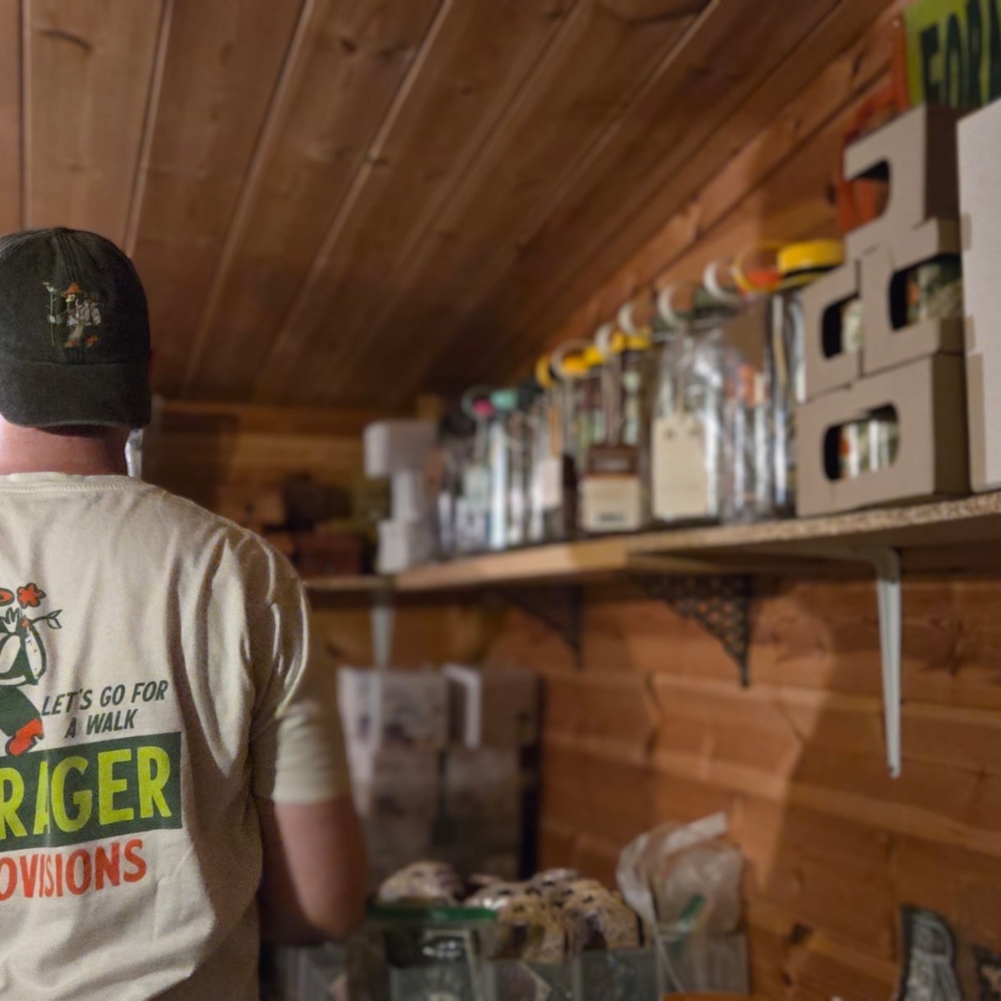 Person in hat and t-shirt stands in rustic wood room, gazing at shelves filled with bottles and containers for storage.