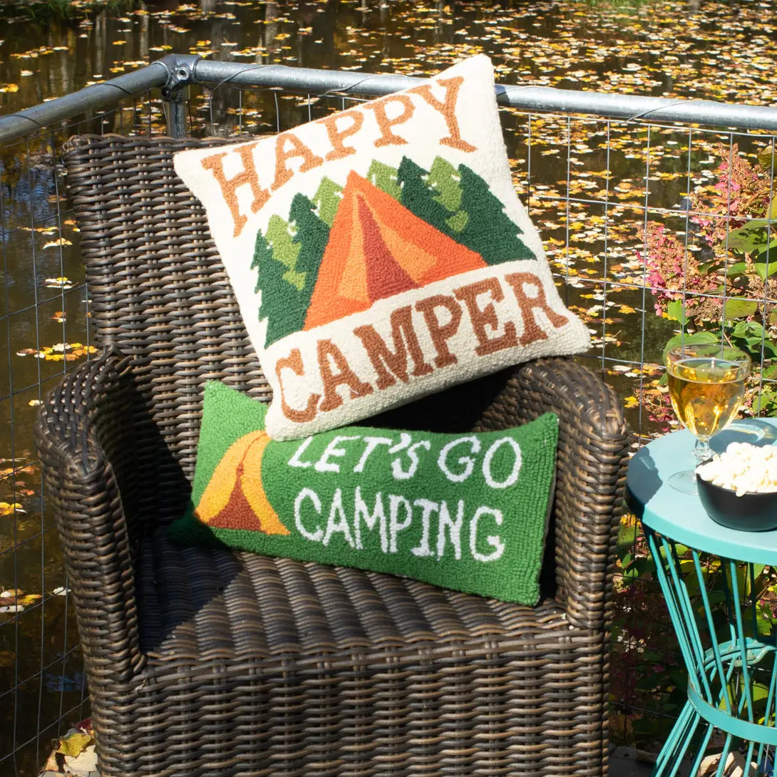 Wicker chair with Peking Happy Camper Wool Hook Cushion by pond, snacks on blue table—relaxing outdoor patio seating scene.