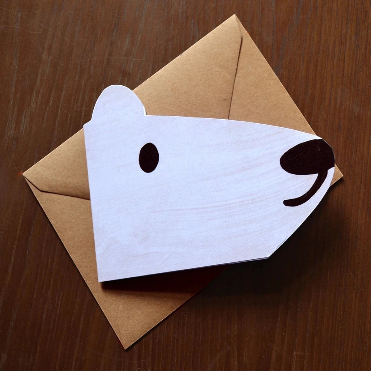 Face of a polar bear shaped card, laid on a brown envelope on a brown wooden table top.