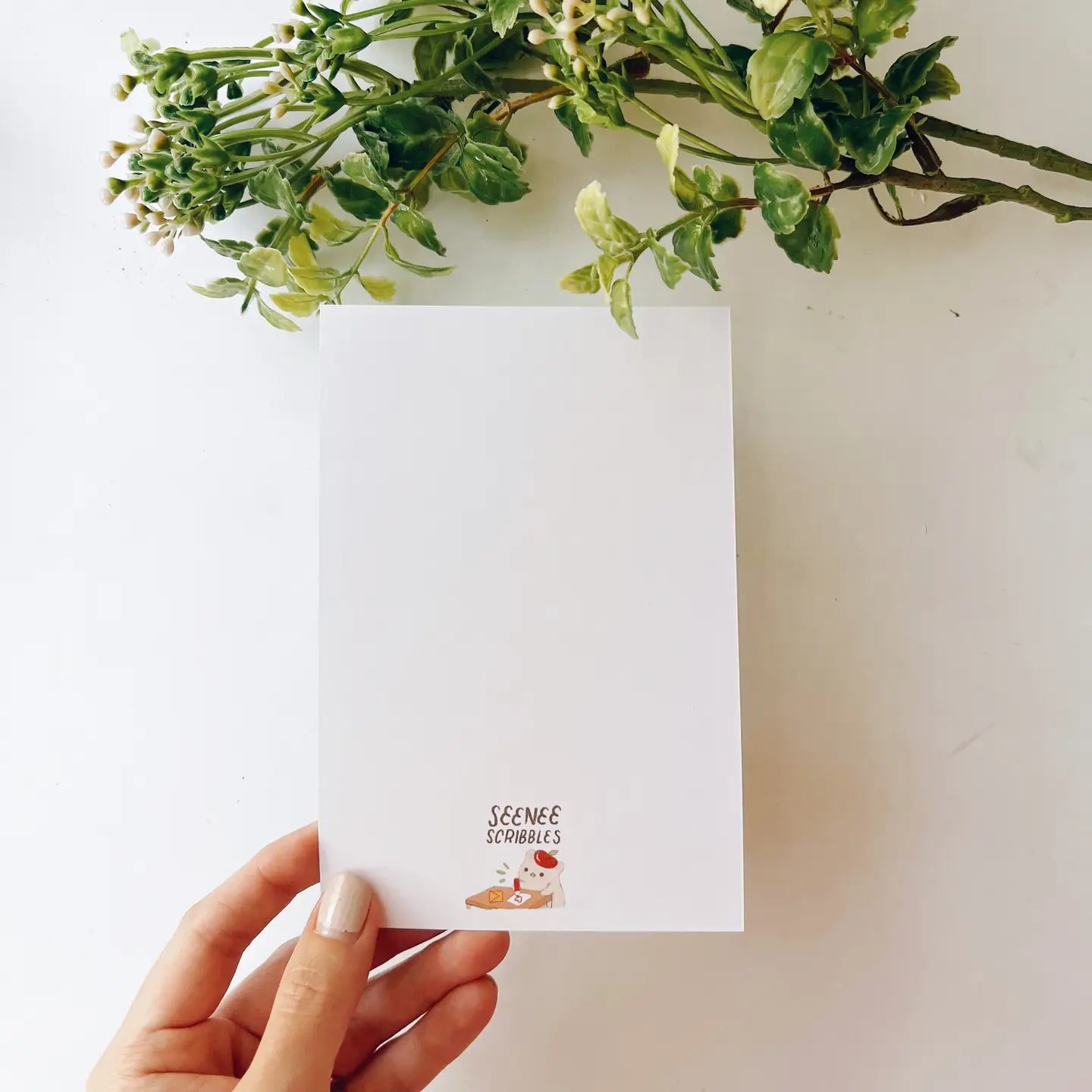 A hand holds the Seenee Scribbles Art Print 4 x 6 - Kiki On A Clear Day, featuring two beautiful watercolor illustrations: a girl at a flowered window (top) and a girl in a field with birds (bottom), evoking scenes from Kiki's Delivery Service.