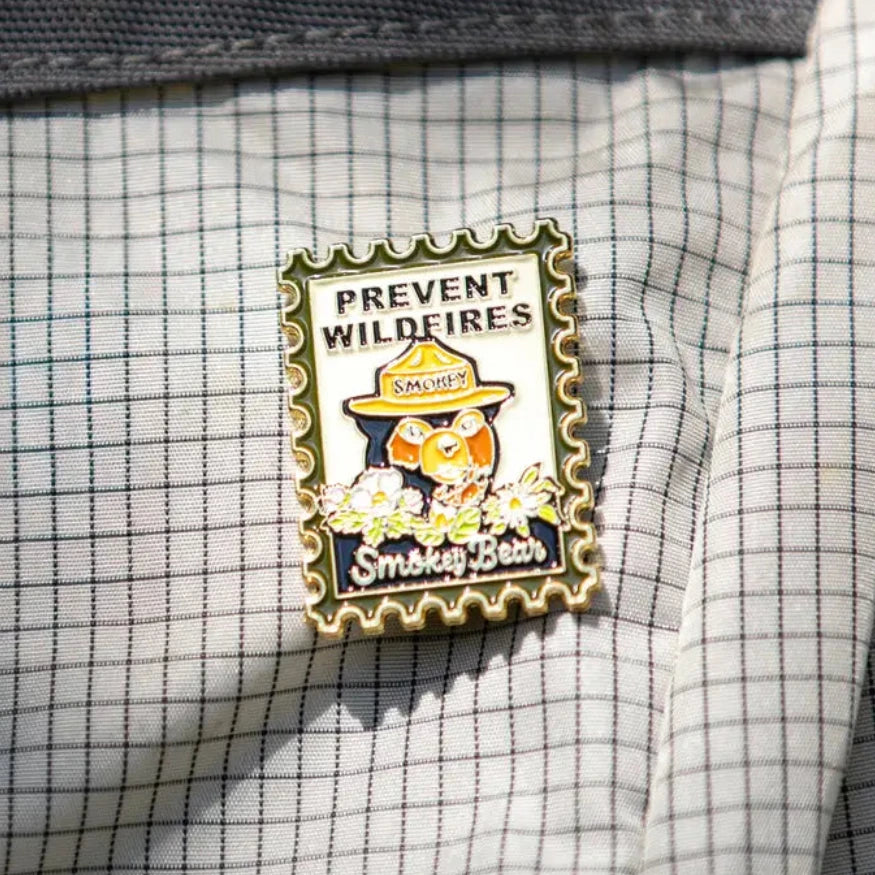 Smokey Bear Stamp Enamel Pin by The Landmark Project on white and gray checkered fabric; perfect forest fire prevention accessory.
