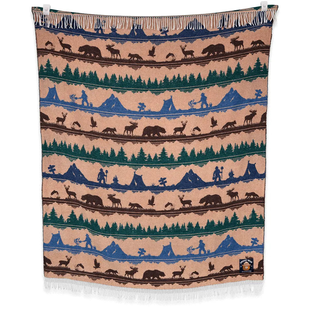 A Sackcloth & Ashes Smokey Bear Blanket - Smokey Dungaree, featuring woven rows with wilderness scenes of trees, mountains, bears, deer, and human figures in neutral tones.