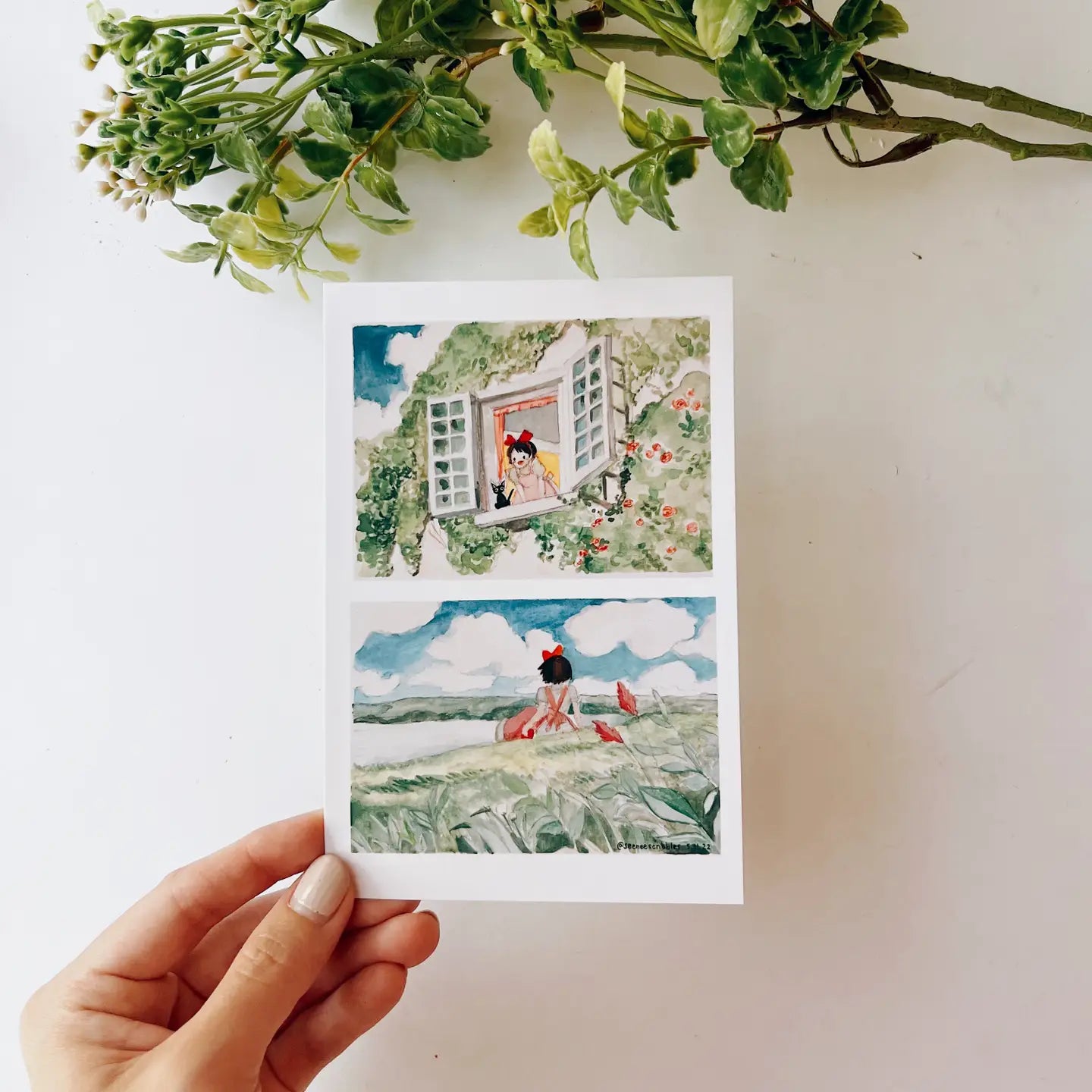 A hand holds the Seenee Scribbles Art Print 4 x 6 - Kiki On A Clear Day, featuring two beautiful watercolor illustrations: a girl at a flowered window (top) and a girl in a field with birds (bottom), evoking scenes from Kiki's Delivery Service.